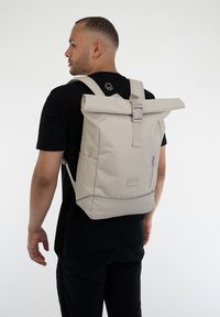 Beige, felhajtható hátizsák, letisztult dizájnnal, szövet anyagból készült, elülső cipzáras zsebbel és állítható pántokkal. Az elején logó található.