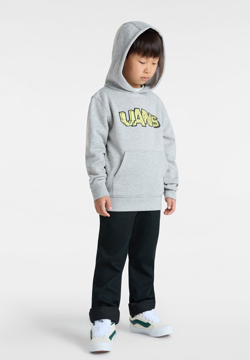 Sweat à capuche gris avec un logo Vans jaune, poche avant et cordon de serrage ; associé à un pantalon sombre et des baskets blanches avec des accents verts.