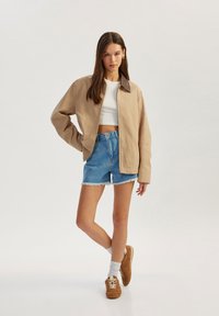Chaqueta beige claro con un cuello marrón, top corto blanco, pantalones cortos de mezclilla azul con dobladillo deshilachado y zapatos casuales marrones combinados con calcetines blancos.