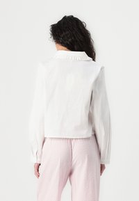 Weißes, cropped Hemd mit Rüschensaum und langen Ärmeln, kombiniert mit strukturierten rosa Hosen. Fokus auf dem Stoff und dem klaren Design.