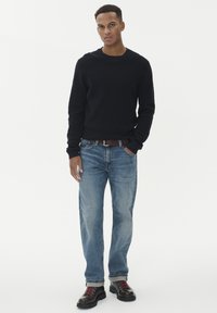 Vyras, dėvintis juodą megztinį, mėlynas džinsus su rudais diržais ir juodais batais su raudonais raišteliais, stovi priešais baltą fono.