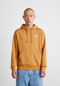 Nike džemperis, sinepes krāsā, izgatavots no mīkstas auduma. Iespējas ietver kangarū durvju kabatu, regulējamu auklu kapuci un mazu baltu logotipu uz krūtīm.