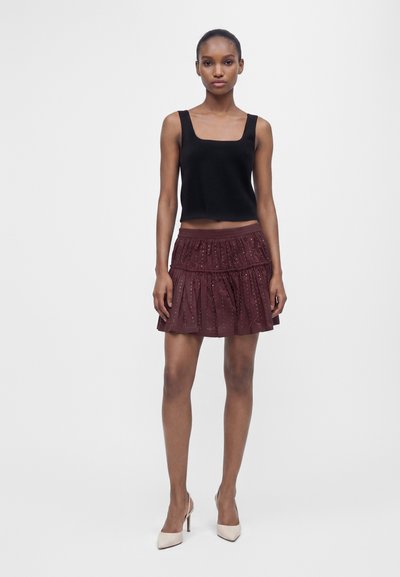Femme portant un crop top noir sans manches, une mini-jupe à volants bordeaux avec des détails œillets et des chaussures blanches à talons hauts, debout devant un fond blanc.