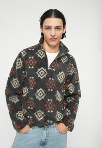 Fleece pulover s polu-zipom, s geometrijskim uzorkom u sivoj, krem i crvenoj boji, s džepom na lijevoj strani prsnog koša.