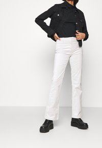 Veste en jean noire avec fermeture à boutons, détails de poches, associée à un pantalon évasé blanc et des bottes à plateforme noires. Texture et design minimalistes.