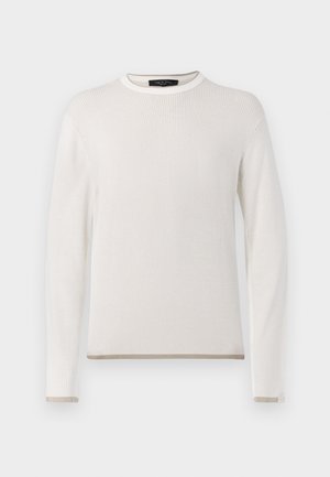 Mies, joka pukeutunut valkoiseen ribbineulottuun pitkähihainen paitaan, tummansinisiin farkkuihin, valkoisiin lenkkareihin ja kantaa mustaa crossbody-laukkua valkoisella taustalla.
