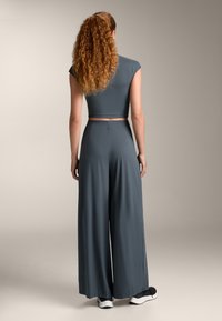 Grijze tweedelige outfit bestaande uit een aansluitend cropped top met kapmouwen en wijde broek, gemaakt van gladde, rekbare stof, in combinatie met zwarte schoenen.