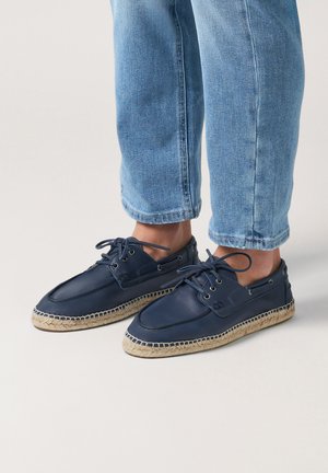 Zapatos de barco azul marino con parte superior de cuero suave, puntera redonda, dos ojales y suela envuelta en yute. Diseño ligero y casual.