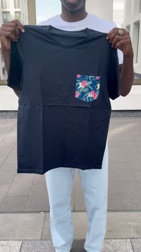 Svart t-skjorte med korte ermer og blå blomsterlomme. Glatt stoff med rund hals og rett kant, holdt av en person som har på seg lyse jeans.