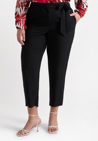 Pantaloni neri a gamba corta con dettaglio a fiocco in vita. Tessuto morbido, vestibilità slim e tasche casual. Abbinati a tacchi bianchi con cinturini.