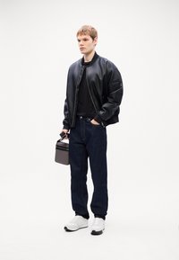 Chaqueta bomber de cuero negra, camiseta negra, vaqueros azul oscuro, zapatillas blancas, sosteniendo un bolso cilíndrico gris con un detalle de cremallera negra.