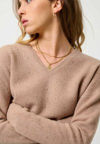 Beżowy sweter z dekoltem w serek o teksturowanym wykończeniu, z drobnymi detalami w postaci kulek, długimi rękawami i prążkowanymi mankietami. Na widoku są dwie warstwowe złote naszyjniki.