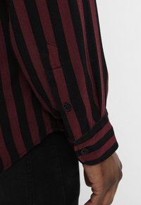 Burgundy and black striped shirt featuring a buttoned cuff, soft texture, and a small front pocket in the sleeve. Minimalistic design.