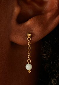 Les boucles d'oreilles en chaîne dorée présentent une petite perle blanche à l'extrémité, suspendue à un clou d'oreille doré rond. Vue rapprochée de l'oreille et des bijoux.