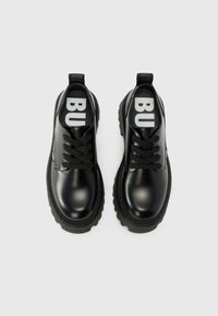 Black leather shoes with a glossy finish, round toe, and thick black sole. Features black laces and visible "BU" branding on the tongue.