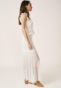 Mujer de perfil vistiendo una blusa sin mangas blanca con volantes y una falda larga plisada, junto con botas de tacón color camel, posando frente a un fondo liso.