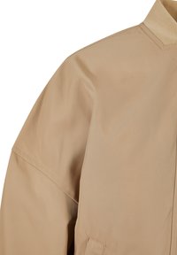 Giacca leggera beige con una texture liscia, vestibilità rilassata e colletto classico. Presenta cuciture minime e un design con una sola tasca.