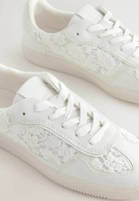 Sneaker bianche con tomaia in pelle texturizzata e dettagli in pizzo sui lati. Presentano lacci tondi e una suola in gomma a profilo basso.