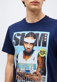 Persona, kas valkā tumši zilu T-kreklu ar Slam žurnāla vāku, kurā redzams Carmelo Anthony Nuggets kreklā un galvas lentā.