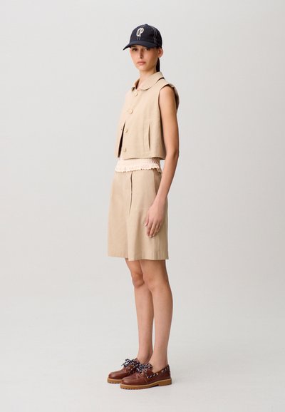 Giovane donna che indossa un gilet beige senza maniche con bottoni, pantaloncini beige della stessa tonalità fino al ginocchio, un berretto blu navy e scarpe marroni stringate, in piedi di profilo.