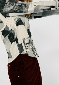 Camicia a maniche lunghe con bottoni in tessuto leggero, caratterizzata da un motivo astratto in bianco e nero, abbinata a pantaloni in corduroy rosso scuro.