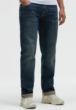 Dark blue denim jeans with straight legs, five-pocket design, and folded cuffs, paired with light-colored sneakers.