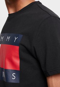 Černé bavlněné tričko s grafickým designem v červené a modré barvě, s nápisem "TOMMY" nad a "S" pod ním, vyvedeným v kontrastním písmu.