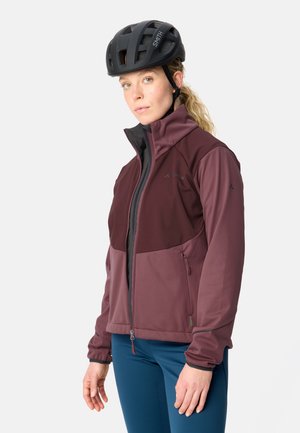 Vrouw met een zwarte fietshelm, een bordeauxrode jas met rits en blauwe broek, staand tegen een effen lichte achtergrond.