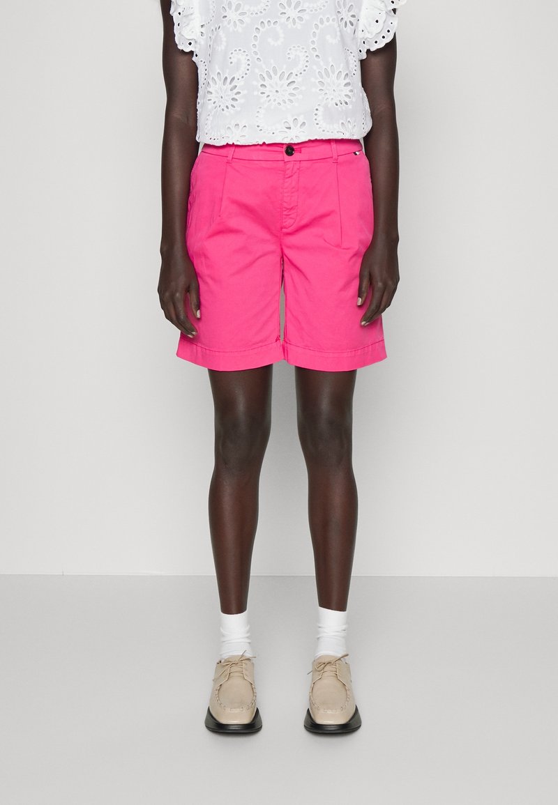 Shorts en coton rose avec poches avant, fermeture par bouton, et coupe décontractée, assortis à un haut blanc brodé et des chaussures beige.