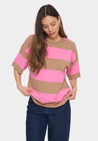 Une femme aux longs cheveux bruns porte une chemise à rayures roses et beige, ample et à manches courtes, ainsi qu'un jean bleu foncé. Elle regarde vers le bas, les mains ajustant l'ourlet de sa chemise.