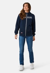 Sweat à zip navy avec le texte blanc "SEASIDE", poches avant, poignets côtelés et ourlet inférieur, assorti à un jean bleu et des baskets blanches.