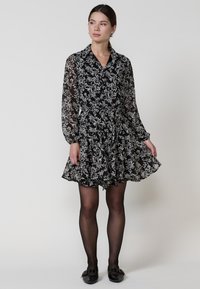Rochie neagră cu flori, cu guler, mâneci lungi din material transparent și talie strânsă. Are o fustă în mai multe straturi și un material ușor, fluid.