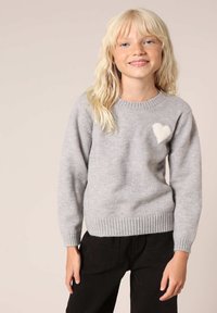 Grijze gebreide trui met een ronde hals en geribde manchetten. Heeft een witte hartaccent op de borst. Gepaard met zwarte broek.