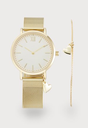 Gouden mesh polshorloge met een ronde witte wijzerplaat, gouden uurmarkeringen en een hart charm. Vergezeld door een dunne gouden kettingarmband met harten.