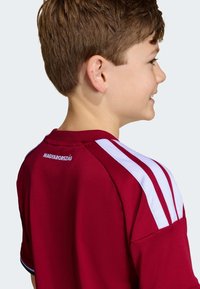 Červený športový dres s bielymi pruhmi na ramenách. Na chrbte text "MAGYARORSZÁG". Hladká tkanina, krátke rukávy, okrúhly výstrih, športový dizajn.