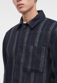 Gros plan d'une personne portant une chemise à carreaux foncés à boutons, avec une poche poitrine frontale et un col, montrant le haut du torse et le cou.