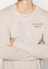 Pitkähihainen, vaaleanbeige paita pehmeästä kankaasta, jossa on pieni "WORLDWIDE" graafinen kuva mustana ja japaninkieliset merkit rinnassa.