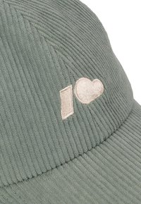 Zielona czapka z sztruksu z wytłoczonym beżowym logo w kształcie litery "I" i serca. Materiał ma pionowe prążki, które nadają projektowi teksturę i głębię.