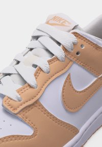 Λευκό και μπεζ αθλητικό παπούτσι Nike σε κοντινή λήψη που δείχνει τα κορδόνια, τις ραμμένες δερμάτινες επιφάνειες και το λογότυπο Nike swoosh στο πλάι.