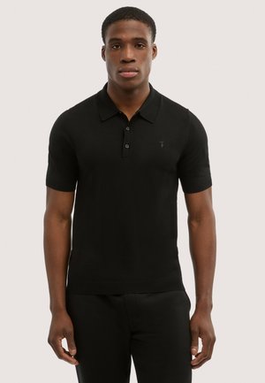 Uomo in piedi di fronte, indossa una polo nera a maniche corte con bottoni e pantaloni neri, su uno sfondo semplice.