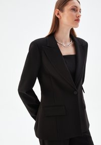 Svart skräddarsydd kavaj med en strukturerad siluett, notchade reverser, enknappsstängning och sidofickor. Slät tygfinish.