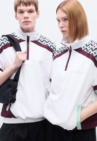 Πουλόβερ-ζακέτα σε λευκό με μαρουν και τιρκουάζ λεπτομέρειες. Διαθέτει φερμουάρ στον γιακά, γεωμετρικά σχέδια στους ώμους και άνετη εφαρμογή.