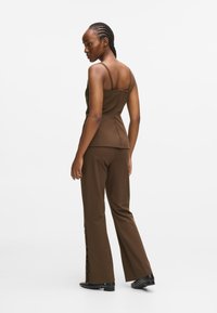 Top maron ajustat cu bretele subțiri și pantaloni maron tailorizați cu accente de butoane pe lateral. Modelul stă cu spatele întors, punând în evidență ținuta.