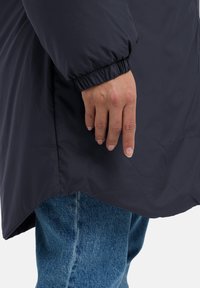 Giacca imbottita blu scuro con una texture liscia, polsini elasticizzati e un orlo posteriore più lungo, abbinata a jeans in denim blu chiaro.