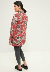 Camicia rossa a maniche lunghe con stampa floreale e animale nera, abbinata a leggings grigi texturizzati e sandali neri a scivolo. Vista posteriore presentata.