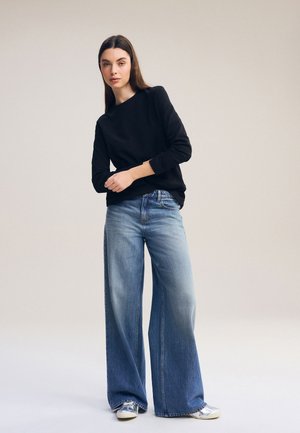 Donna in piedi che indossa un top nero a maniche lunghe, jeans blu a gamba larga e sneakers argentate su uno sfondo beige uniforme.