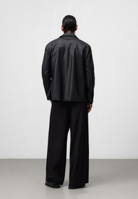 Blazer in pelle nera con colletto strutturato, vestibilità dritta e dettagli minimi. Indossato con pantaloni neri larghi e scarpe basse.
