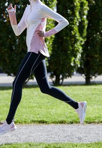 Lehký růžový a bílý sportovní top s vysokým límcem a otvory na palce, doplněný černými legínami s bílými bočními pruhy. Sportovní obuv.