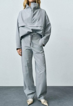 Veste grise surdimensionnée à demi-zip avec des manches longues, associée à un pantalon large assorti et des chaussures pointues de couleur crème. Texture de tissu lisse.