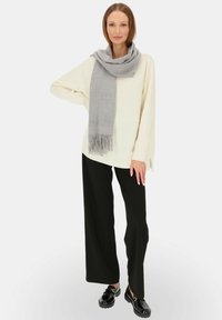 Kremowy sweter, szary szalik z frędzlami, czarne spodnie o szerokich nogawkach oraz czarne buty. Szalik jest elegancko zarzucony na szyi.